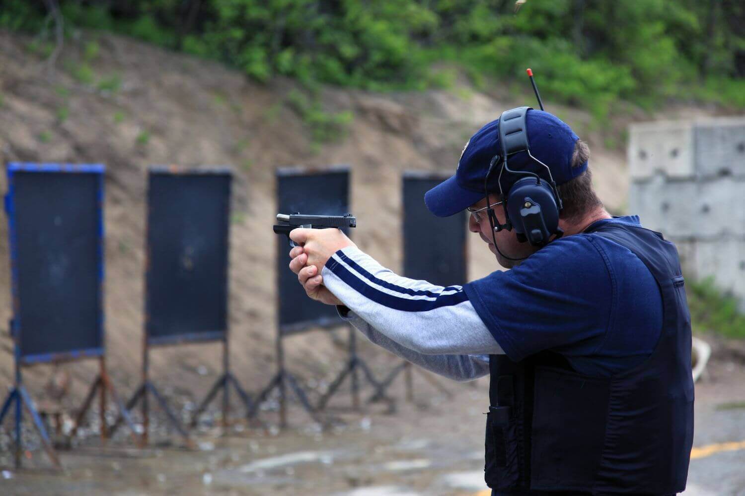 Man shooting a 9mm handgun at an outdoor shooting range. He's wearing protective gear for his ears and eyes while aiming at his target