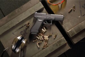 Black pistol with multiple loose bullets and a set of protective earmuffs on a crate