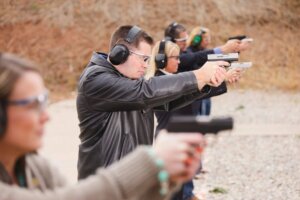 A group of men and women wearing protective gear are firing various pistols at an outdoor shooting range