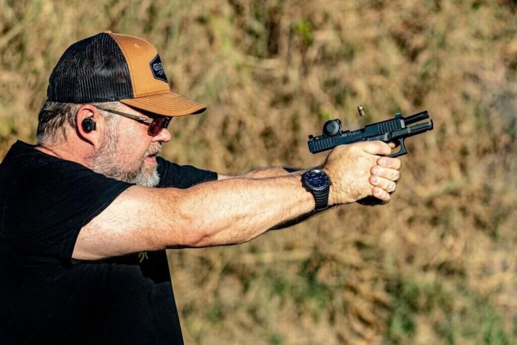 Man in a cap and protective eyewear aiming a handgun outdoors, with a shell casing visible mid-air