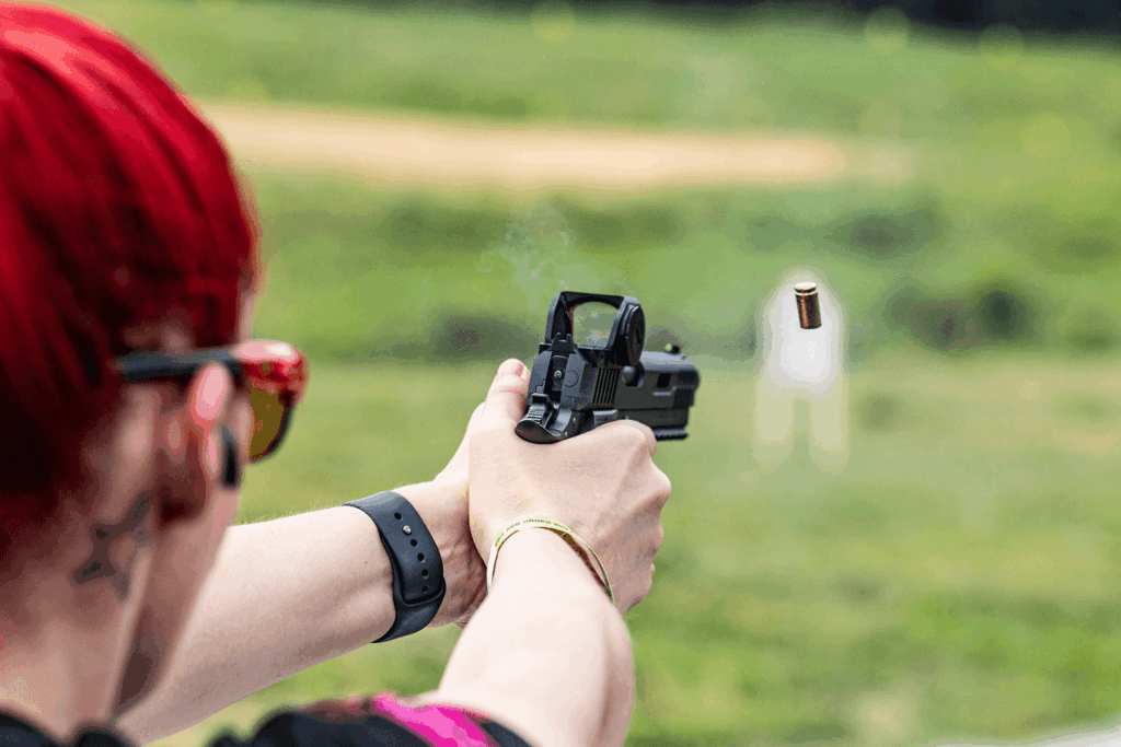 Woman with red hair firing a pistol with an optic mounted at an outdoor shooting range - JSD Supply | Premier Gun Parts And Accessories Woman with red hair firing a pistol with an optic mounted at an outdoor shooting range