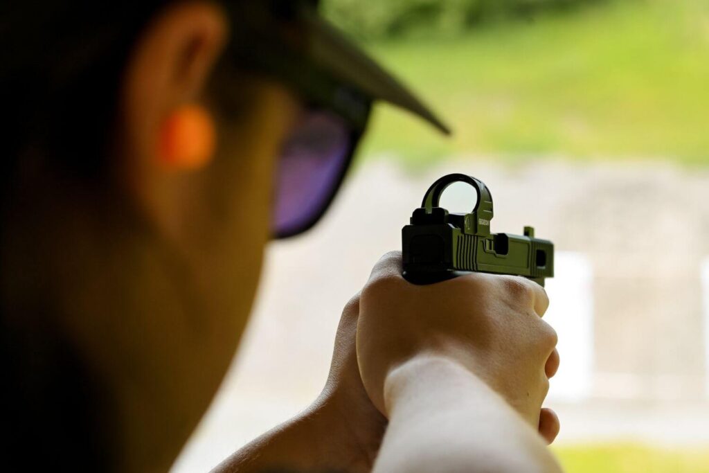 View behind a man aiming a firearm using a mounted red dot optic